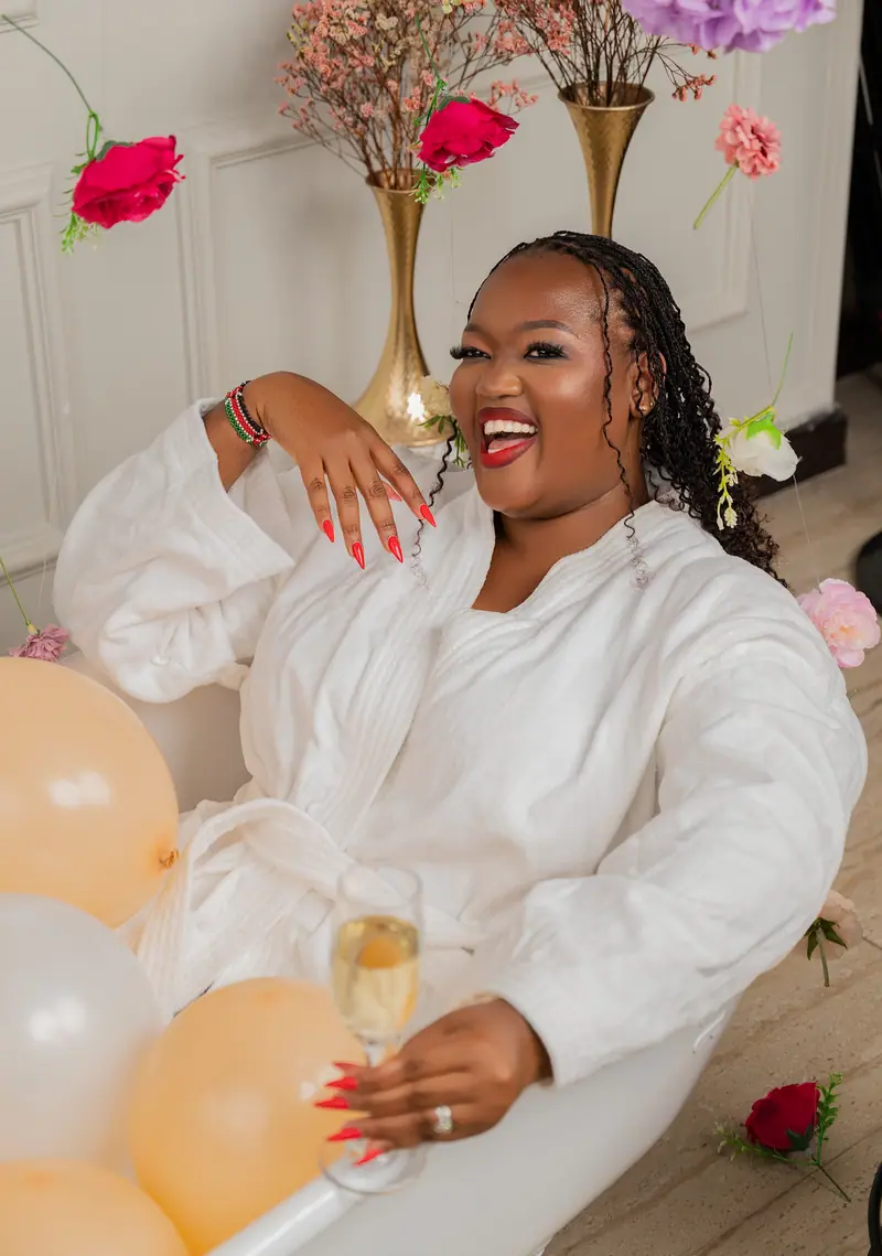 Woman laughing in a white robe, holding champagne, among balloons and flowers